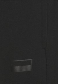 Sort stof med en glat overflade og et rektangulært sort labelskilt. Har en sidesøm og et minimalistisk design.
