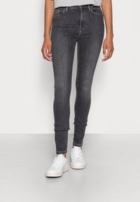 Mujer vestida con pantalones ajustados gris oscuro y zapatillas blancas, de pie sobre un suelo gris claro contra una pared blanca.