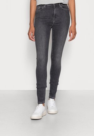 Mujer vestida con pantalones ajustados gris oscuro y zapatillas blancas, de pie sobre un suelo gris claro contra una pared blanca.