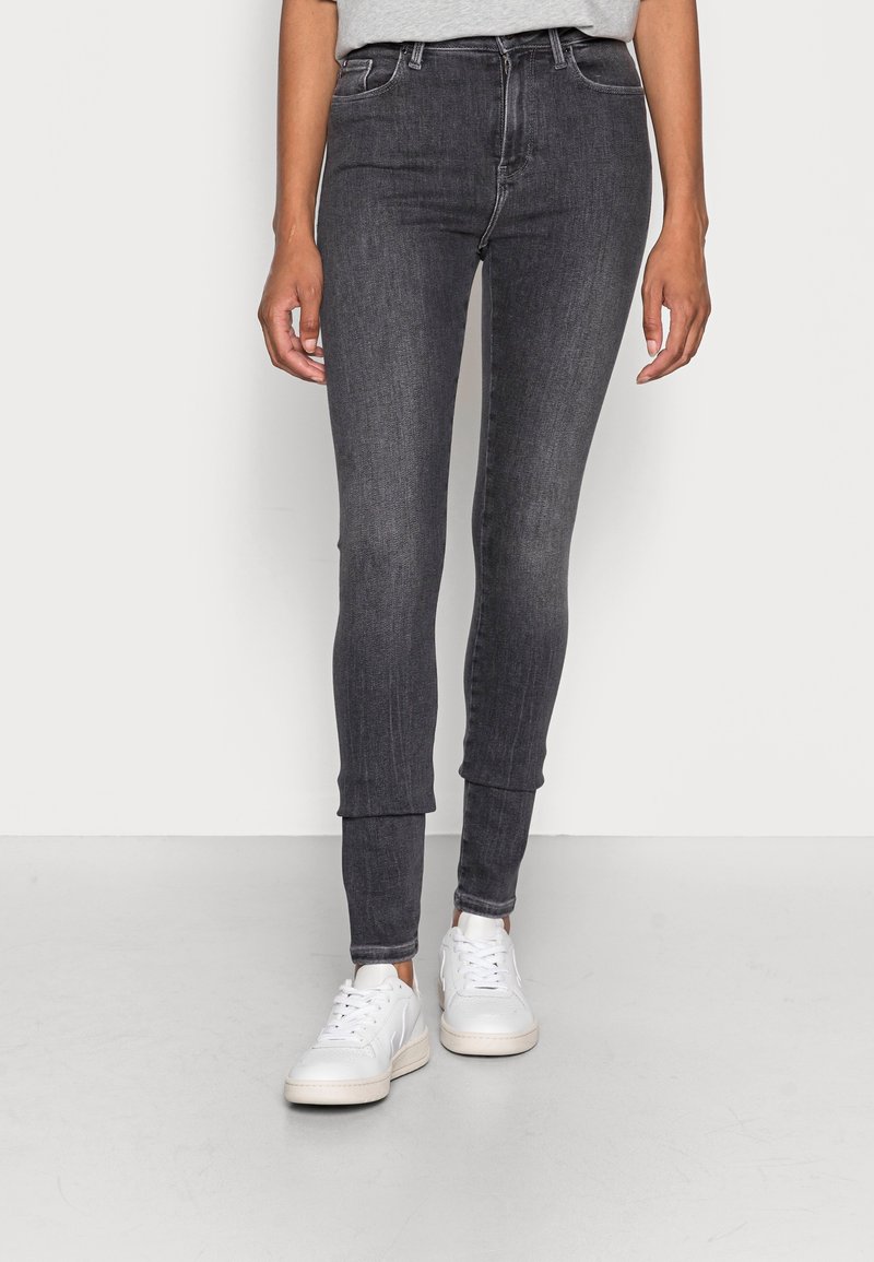 Mujer vestida con pantalones ajustados gris oscuro y zapatillas blancas, de pie sobre un suelo gris claro contra una pared blanca.