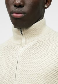 Maglione beige a mezza zip in maglia con motivo testurizzato, colletto rialzato e dettaglio della zip in argento. Vista ravvicinata che mette in evidenza il tessuto e il design.