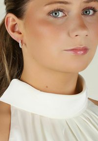 Vit satinöverdel med hög krage och veckad design; modellen bär en glittrande hoop-örhänge med diamanter, slät hud.