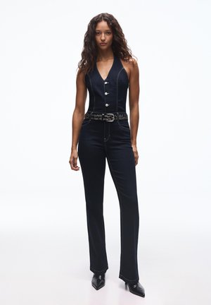 Kvinde med krøllet hår iført en tætsiddende, ærmeløs sort jumpsuit med knapper, et nittebælte og sorte spidse sko, stående mod en hvid baggrund.