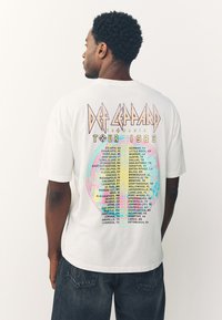 White cotton t-shirt featuring a colorful graphic design on the back with "Def Leppard Tour 1983" text and a detailed list of cities.