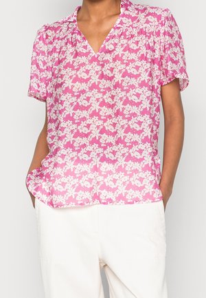 Osoba ubrana w różową bluzkę z krótkim rękawem z białym kwiatowym wzorem i białe spodnie, dłonie w kieszeniach, na jednolitym tle.