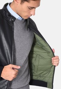 Giacca in pelle nera con fodera trapuntata verde, dotata di chiusura con zip e polsini elastici. Indossata sopra un maglione grigio.