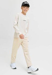 Beige langwerpige shirt met "JJ Studio" logo, bijpassende lichte broek en zwarte pet, gecombineerd met witte sportschoenen. Soepele stof textuur.