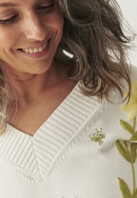 Biały sweter z dzianiny z żebrowanym dekoltem w kształcie V, ozdobiony zieloną i żółtą haftowaną florystyką oraz małym motywem pszczoły na ramieniu.