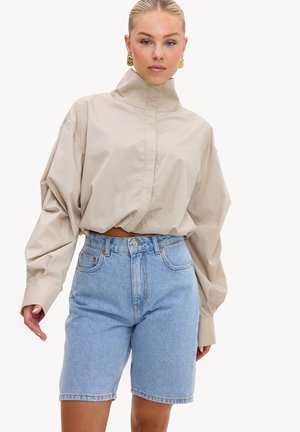 Femme aux cheveux blonds portant un chemisier beige à col haut et manches longues et un short en jean bleu clair taille haute, debout sur un fond blanc.