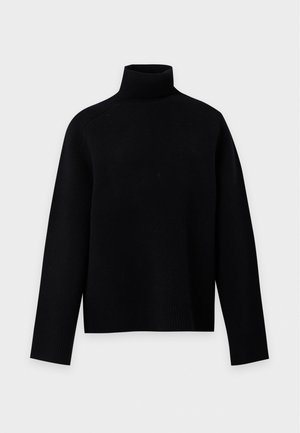 Femme aux longs cheveux foncés portant un pull à col roulé noir, une longue jupe en cuir noire et des bottines noires à talons, debout sur un fond blanc.