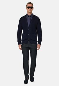 Cardigan in maglia blu navy con bottoni, indossato sopra una camicia scura con bottoni e pantaloni scuri a sigaretta. Il modello indossa occhiali da sole neri e scarpe nere.