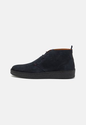 Botines de ante azul oscuro con una suela de goma negra texturizada, un diseño sencillo de cordones y un interior marrón en contraste.