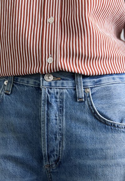 Ριγέ πουκάμισο σε κόκκινο και λευκό χρώμα πάνω από ξεθωριασμένο μπλε τζιν παντελόνι, με μεταλλικό κουμπί και τσέπες με διακριτική φθορά.