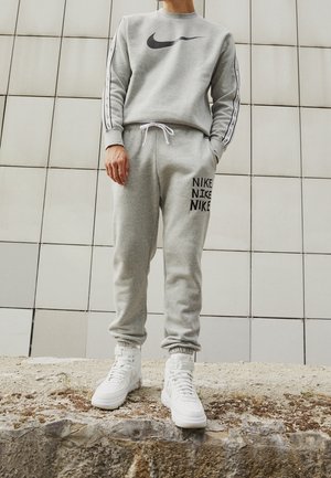 Person iført gråt Nike træningssæt og hvide Nike høje sneakers, stående på en rå stensurface mod en flisebelagt væg i baggrunden.