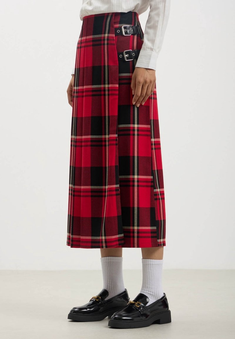 Femme portant une jupe midi en carreaux rouge et noir avec deux sangles noires à boucle, des chaussettes blanches, des mocassins noirs et une chemise blanche à manches longues.