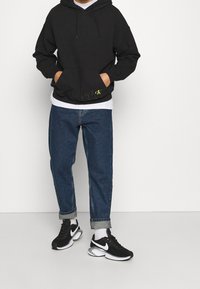 Sudadera negra con un bolsillo frontal y un logotipo visible en amarillo, camiseta blanca debajo, jeans de denim azules y zapatillas Nike negras con acentos blancos.