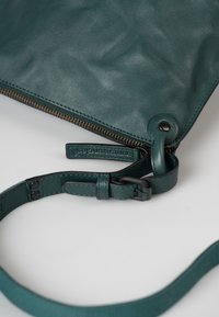 Borsa a tracolla verde in pelle con superficie texturizzata, zip in bronzo e tracolla removibile. Presenta un piccolo etichetta con il marchio vicino al giunto della tracolla.