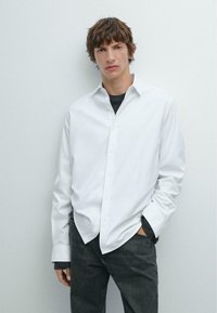 Weißes Langarm-Hemd mit Kragen, kombiniert mit einem dunkelgrauen T-Shirt und dunklen Jeans. Glatte Textur, minimalistisches Design.