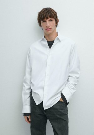 Weißes Langarm-Hemd mit Kragen, kombiniert mit einem dunkelgrauen T-Shirt und dunklen Jeans. Glatte Textur, minimalistisches Design.