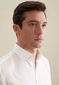 Biała koszula z guzikami, o sztywnym kołnierzyku i gładkiej tkaninie. Projekt charakteryzuje się minimalistyczną estetyką, bez wzorów czy akcentów.
