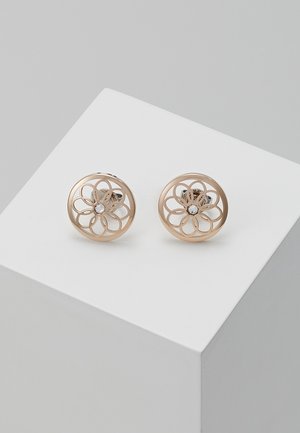 Boucles d'oreilles rondes en ton or avec un motif floral ajouré et un rhinestone clair central, présentées sur un piédestal géométrique blanc.