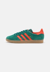 Gröna mocka Adidas Gazelle-sneakers med orangea ränder, hälflik och sula av gummi, visade från sidan på en vit bakgrund.