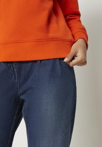 Pomarańczowa bluza z prążkowanym ściągaczem u dołu, zestawiona z ciemnoniebieskimi spodniami dżinsowymi z elastycznym pasem i subtelną fakturą.