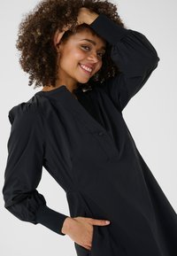 Besijuokianti moteris su garbanotais plaukais, dėvinti ilgomis rankovėmis juodą suknelę, viena ranka laikosi už galvos, kita ranka kišenėje, priešais paprastą foną.