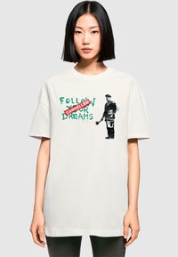 Oversized white cotton t-shirt featuring green text "FOLLOW YOUR DREAMS" with "CANCELLED" in red, and a graphic of a man holding a paintbrush.