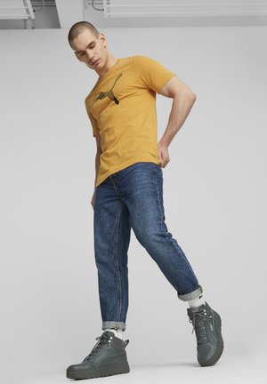 Camiseta amarilla de manga corta con un diseño gráfico negro, combinada con jeans de mezclilla azules y zapatillas altas grises.