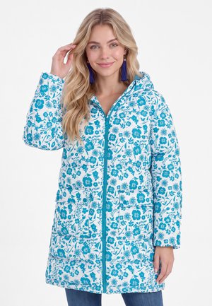 Blonde Frau trägt eine weiße und blau gemusterte Kapuzen-Steppjacke mit Blumenmuster, Vorderentaschen und blaue Quasten-Ohrringe.