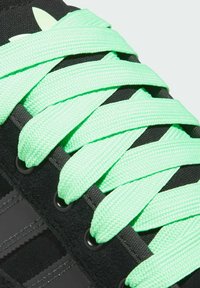 Černá teniska se širokými černými pruhy a jasně zelenými tkaničkami propletenými kulatými očky, s texturovaným povrchem. 