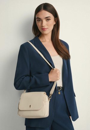 Kobieta w granatowym garniturze trzyma beżową skórzaną torebkę na ramię ze złotymi elementami, stojąc na jednolitym jasnym tle.