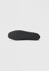 Sole di una scarpa nera senza lacci con scanalature orizzontali per la presa, mostrata dal fondo su uno sfondo bianco semplice.