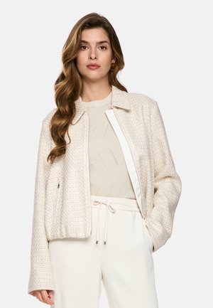 Frau mit langen, welligen Haaren trägt eine strukturierte, cremefarbene Jacke über einem hellen Strickpullover und hoch taillierte weiße Hose mit Kordelzug.