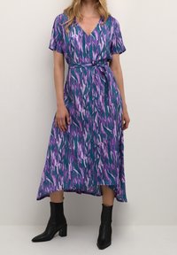 Robe longue avec motif abstrait violet, vert et blanc ; manches courtes, ceinture à nouer à la taille, décolleté en V, avec détails boutonnés. Portée avec des bottines noires.