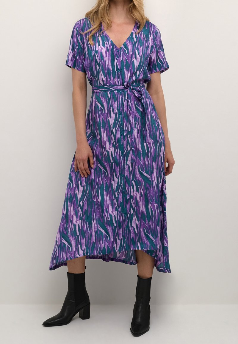 Robe longue avec motif abstrait violet, vert et blanc ; manches courtes, ceinture à nouer à la taille, décolleté en V, avec détails boutonnés. Portée avec des bottines noires.