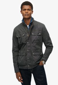 Grå letvægtsjakke med ståkrave, fire frontlommer og knaplukning; parret med en denimskjorte og mørke jeans.