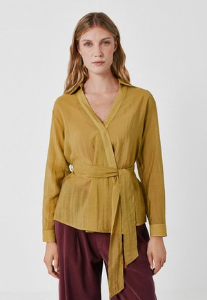 Donna con indosso una camicetta avvolgente giallo senape con cintura in vita e maniche lunghe, abbinata a pantaloni larghi color bordeaux, in piedi davanti a uno sfondo semplice.