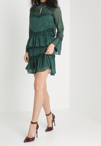 Grön chiffongklänning med lager av volanger och spetsdetaljer. Långa ärmar och hög halsringning. Bärs med bourgognefärgade klackskor.