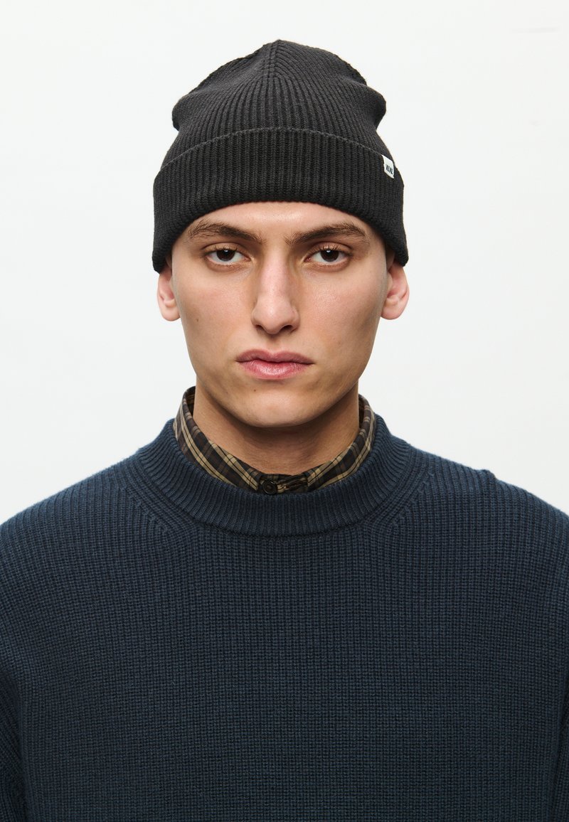 Black ribbed beanie hat, featuring a turned-up cuff. The model wears a dark blue textured sweater over a plaid shirt.