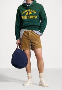 Person som bär en grön hoodie med texten "North East Cross Country", randig skjorta, bruna kordflöjelshorts, beige strumpor, vita sneakers och håller en marinblå duffelväska.