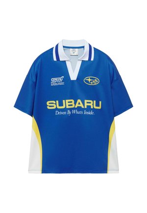 Młody mężczyzna stoi ze złożonymi rękami za plecami, ubrany w niebieską koszulkę z logo Subaru, ciemne krótkie spodenki do kolan, białe skarpetki i beżowe trampki.