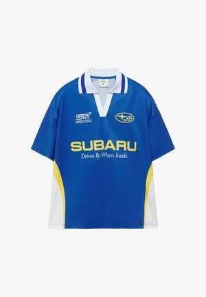 Giovane uomo in piedi con le mani dietro la schiena, indossa una maglietta blu con il marchio Subaru, pantaloncini scuri fino al ginocchio, calzini bianchi e scarpe da ginnastica beige.