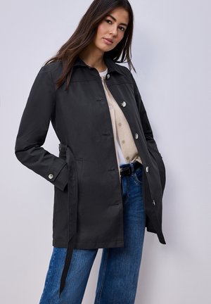 Nainen pukeutuneena mustaan napitettuun takkiin vyöllä, beige neuletakki, valkoinen paita ja siniset farkut, seisomassa vaaleanharmaan taustan edessä.