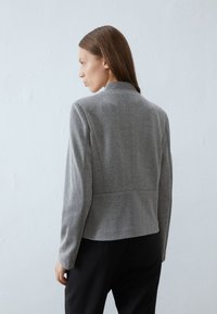 Giacca grigia con motivo a spina di pesce, dal taglio sartoriale, leggermente accorciata e con colletto alto, abbinata a pantaloni neri.