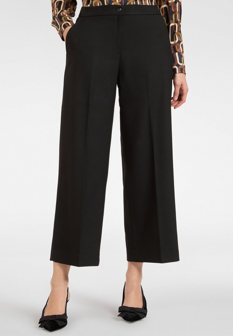 Czarne szerokie spodnie o gładkiej fakturze, z przednimi kieszeniami i zapięciem na guzik, zestawione z czarnymi szpilkami z ostro zakończonym noskiem.