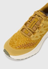 Scarpa sportiva gialla con tomaia in maglia strutturata, lacci piatti e suola ammortizzata. Dettagli notevoli includono un design a motivo e accenti decorativi.