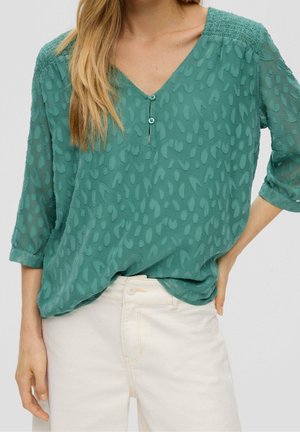 Vrouw draagt teal gestructureerde blouse met knopen en driekwart mouwen, gecombineerd met witte broek met hoge taille, hand op heup.