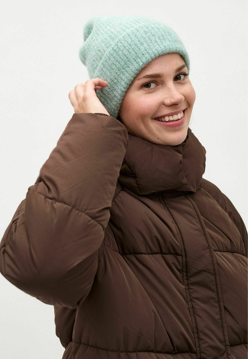 Chaqueta de plumas marrón con un cuello alto, con un diseño acolchado. Gorro de punto de color azul claro y textura acanalada. Persona sonriendo mientras se ajusta el gorro.